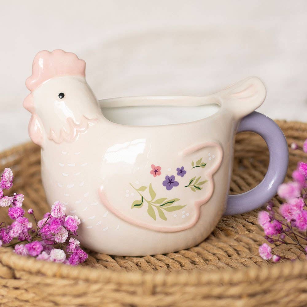 Ceramic chicken mug with floral design on a woven surface with pink flowers.