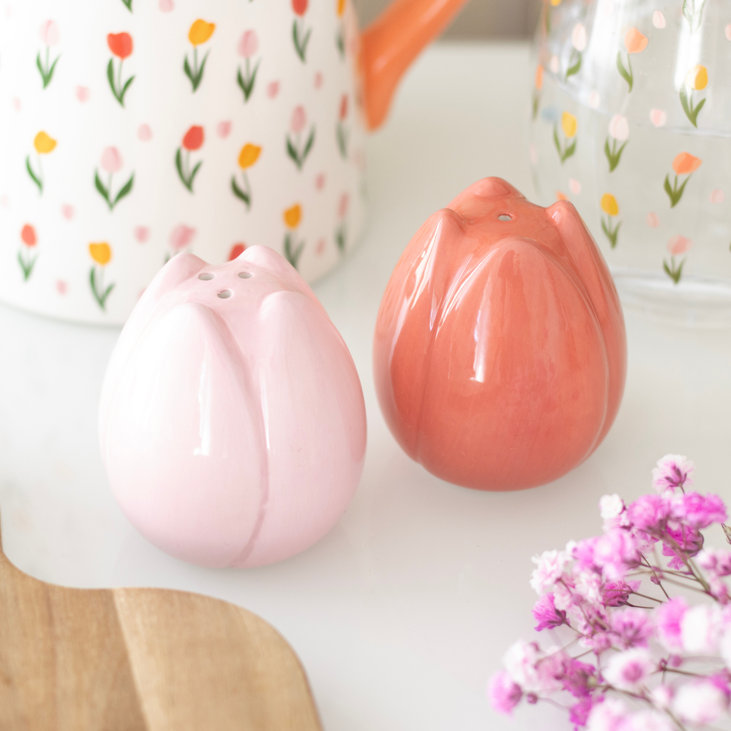 Two pink ceramic tulip-shaped items on a white surface with floral-patterned mugs in the background.