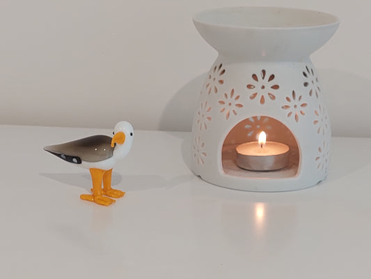 White ceramic oil burner with a lit candle and a small bird figurine on a white surface.